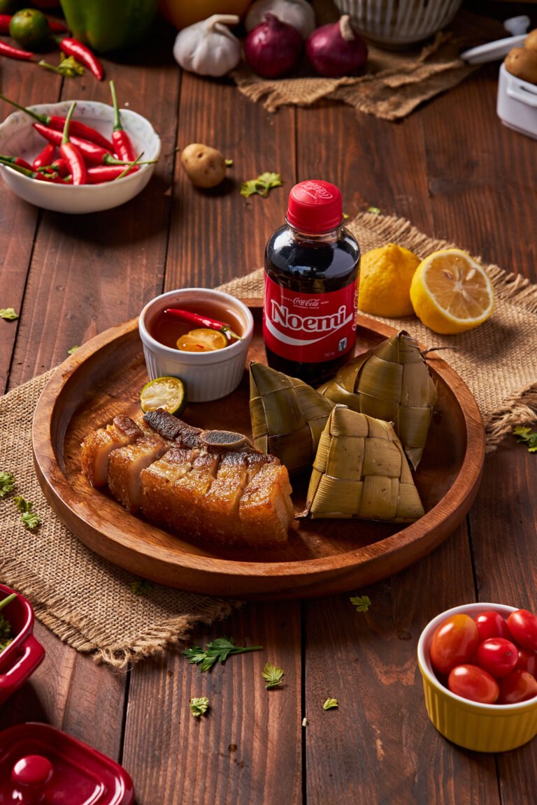 Appetizing photo of deep-fried, bone-in pork belly with extremely crispy skin, accompanied by a savory dipping sauce and traditional hanging rice packets (pusô).