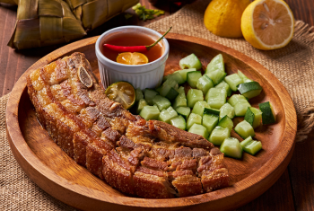 Appetizing photo of deep-fried, bone-in pork belly with extremely crispy skin, accompanied by a savory dipping sauce and traditional hanging rice packets (pusô).