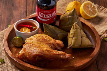 A Filipino dish featuring a piece of fried chicken served over rice, two triangular packets of woven rice (Puso), and a dipping sauce with chili and calamansi, all on a wooden plate. A bottle of personalized Coca-Cola is in the background.