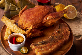 A wooden plate featuring a batter_fried whole chicken, a slab of crispy pork belly, spring rolls, and a dipping sauce with calamansi.